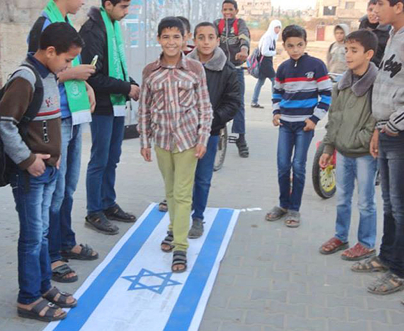 »Al-Kutla-al-Islamiya«, die für Schüler und Studenten zuständige »Bildungsabteilung« der Hamas, feiert in der UNRWA-geführten Yabna-Mittelschule in Rafah einen »Sieg« über Israel, indem sie Schüler unter anderem auf der israelischen Fahne herumtrampeln lässt. Rafah, 1. Dezember 2013